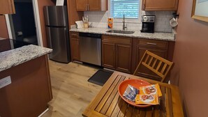 A compact kitchen with wooden cabinets, a stainless steel refrigerator, a dishwasher, and a window with blinds. - Barefoot Cottage A Quiet Coastal Retreat (annapolis)