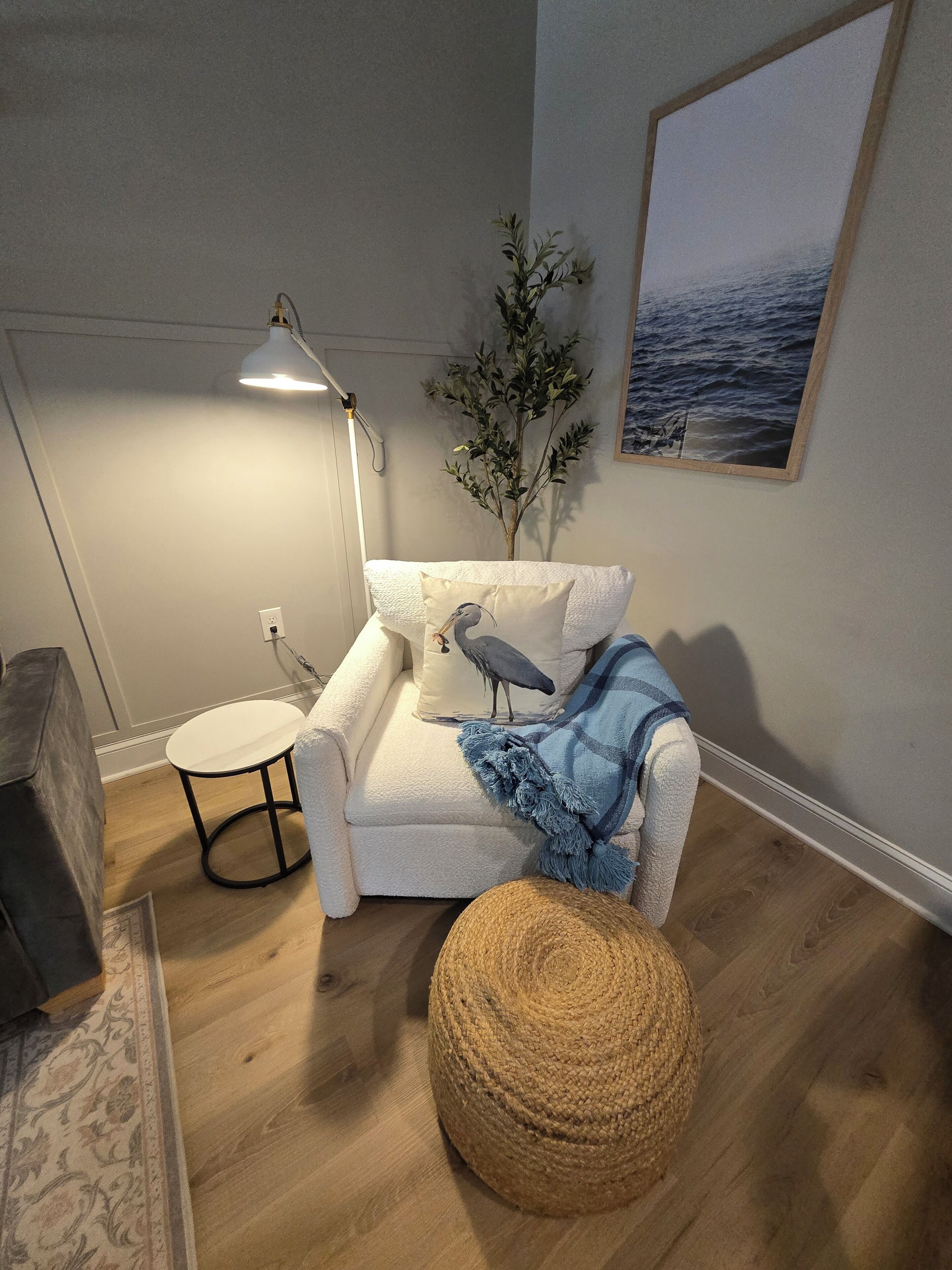 A cozy living room with a white armchair, a small round table, a lamp, a plant, and a framed picture on the wall.
