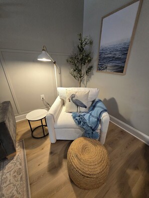 A cozy living room with a white armchair, a small round table, a lamp, a plant, and a framed picture on the wall.