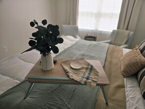 A neatly arranged bedroom with a bed, a bedside table with a vase and a remote control, and a window with curtains.
