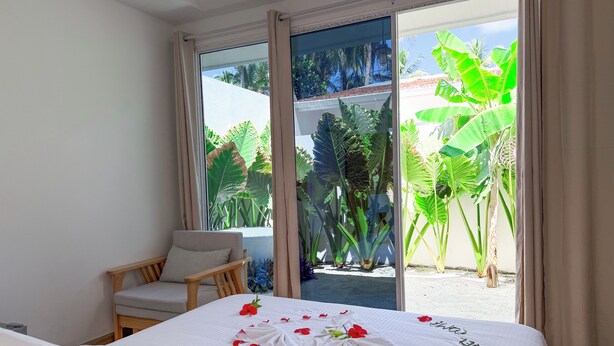 A hotel room with a bed, a chair, and a view of tropical plants through a glass door. - Tropicana Villas - Thoddoo (Thoddoo)