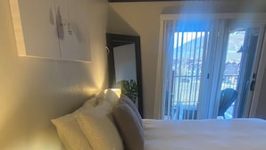 Quarto de hotel com cama, mesa de cabeceira com abajur, mesinha de apoio com caixa de lenços e vista para o exterior por uma porta de correr.