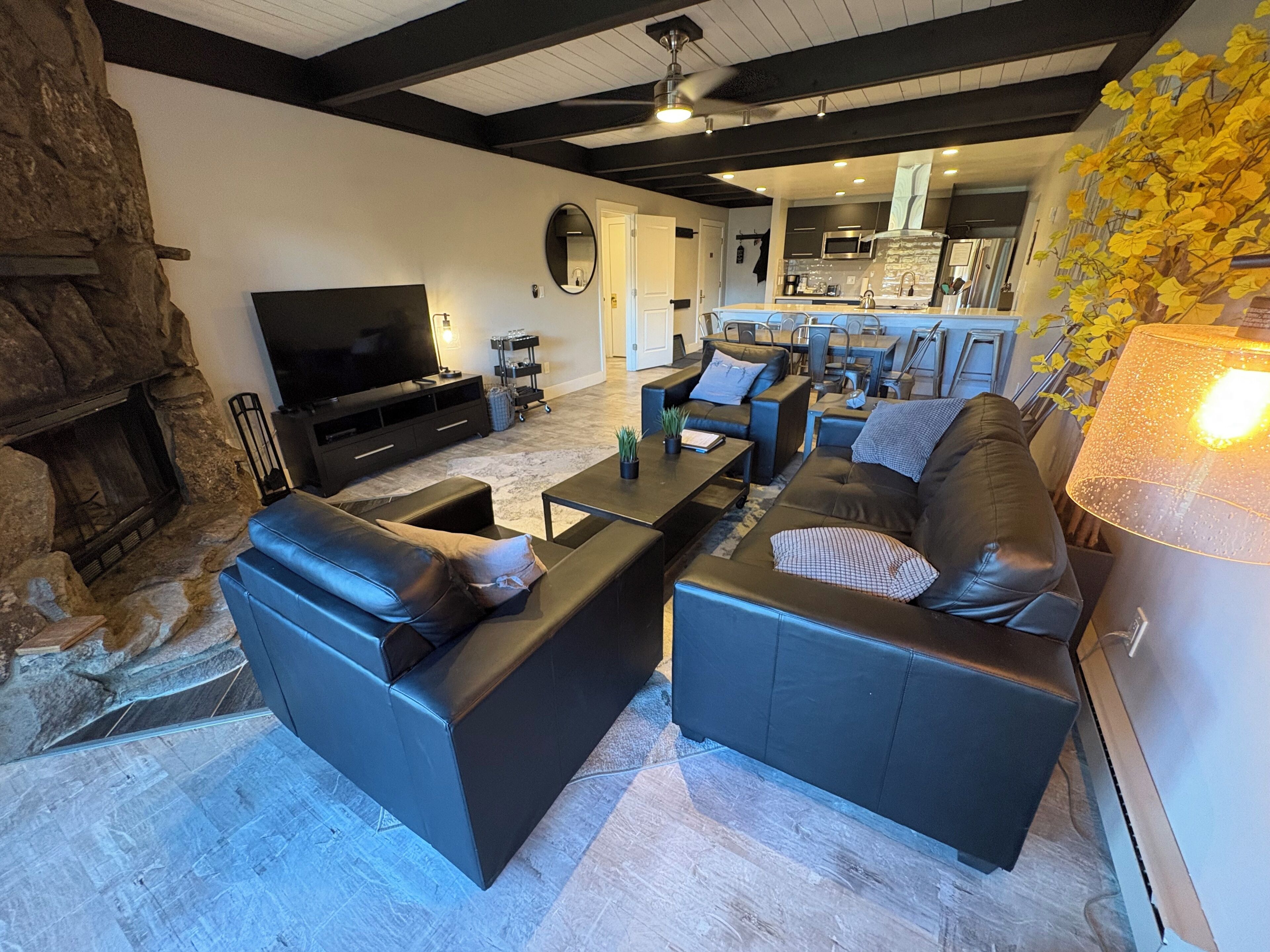 Ein modernes Wohnzimmer mit Kamin aus Naturstein, schwarzen Ledersofas, einem Flachbildfernseher und einer Küche im Hintergrund.