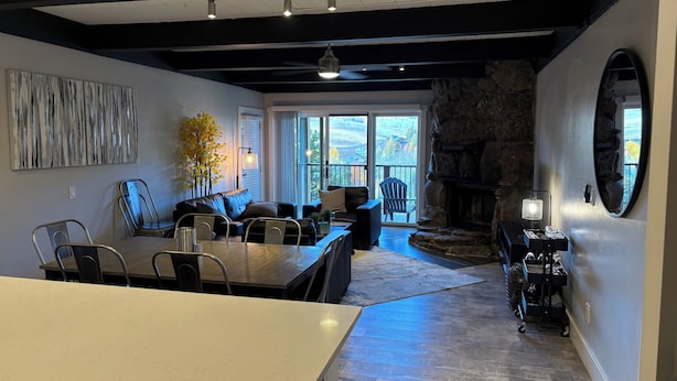 Uma sala de estar moderna com lareira de pedra, canto de jantar com mesa e cadeiras, e uma grande janela com vista para o exterior.