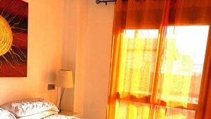 A neatly made bed with white bedding and two rolled towels, a painting on the wall, and a window with orange curtains. - Sea sunrise (Guardamar del Segura)
