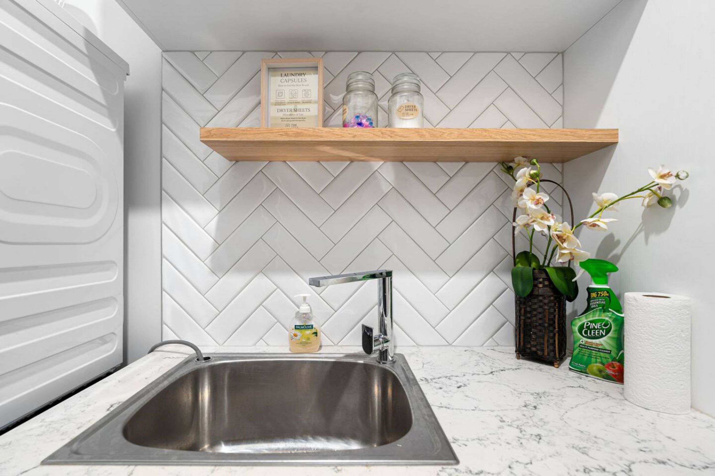 A kitchen with a stainless steel sink, a wooden shelf with laundry products, and a potted orchid.