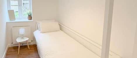 A small, single-bed room with a wooden floor, a window with a view, a radiator, and a small table with a lamp.
