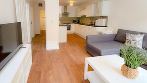 Ein modernes Wohnzimmer mit einer grauen Couch, einem weißen Couchtisch und einem Flachbildfernseher. Im Hintergrund ist eine Küche mit weißen Schränken sowie ein Essbereich mit Tisch und Stühlen erkennbar.