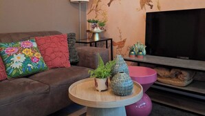 Ein Wohnzimmer mit einer Couch, einem runden Holztisch, einem pinken Akzentstuhl, einem Fernseher und einer Wand mit einem Vogelgemälde.