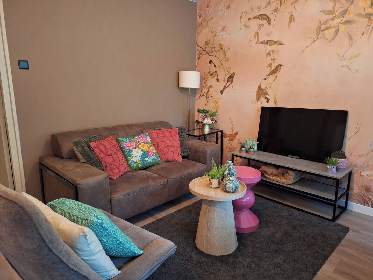 A living room with a brown sofa, a smaller teal sofa, a coffee table, a TV stand with a flat-screen TV, and a floral wallpaper.