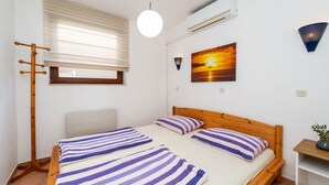 Ein Schlafzimmer mit einem hölzernen Bett, gestreifte Kissen, einem Fenster mit Jalousien, einer Wand-Klimaanlage und einem Gemälde vom Sonnenuntergang.