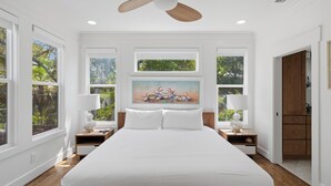 Une chambre à coucher avec un lit, deux tables de chevet, un ventilateur de plafond, un tableau et de grandes fenêtres.