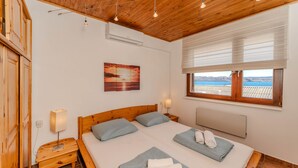 Un dormitorio con techo de madera, una cama con dos almohadas, una mesita de noche, un aire acondicionado de pared, una ventana con vistas y un cuadro de atardecer.