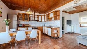 Cocina moderna con armarios de madera, zona de comedor con sillas blancas, televisor y ventana con persianas.