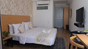 Una camera d'albergo con un letto rifatto, una scrivania con una sedia, un televisore e una porta che dà su un'altra stanza.