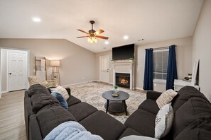 A living room with a fireplace, a ceiling fan, and a large sectional sofa. - Downtown Oasis • Hot Tub + Game Room Fun - Cozy (Bentonville)