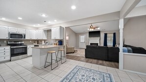 Une cuisine moderne avec des meubles blancs, un îlot central doté de tabourets, un four à micro-ondes et un four traditionnel. Le salon est agrémenté d’un canapé de couleur foncée et d’un ventilateur de plafond.