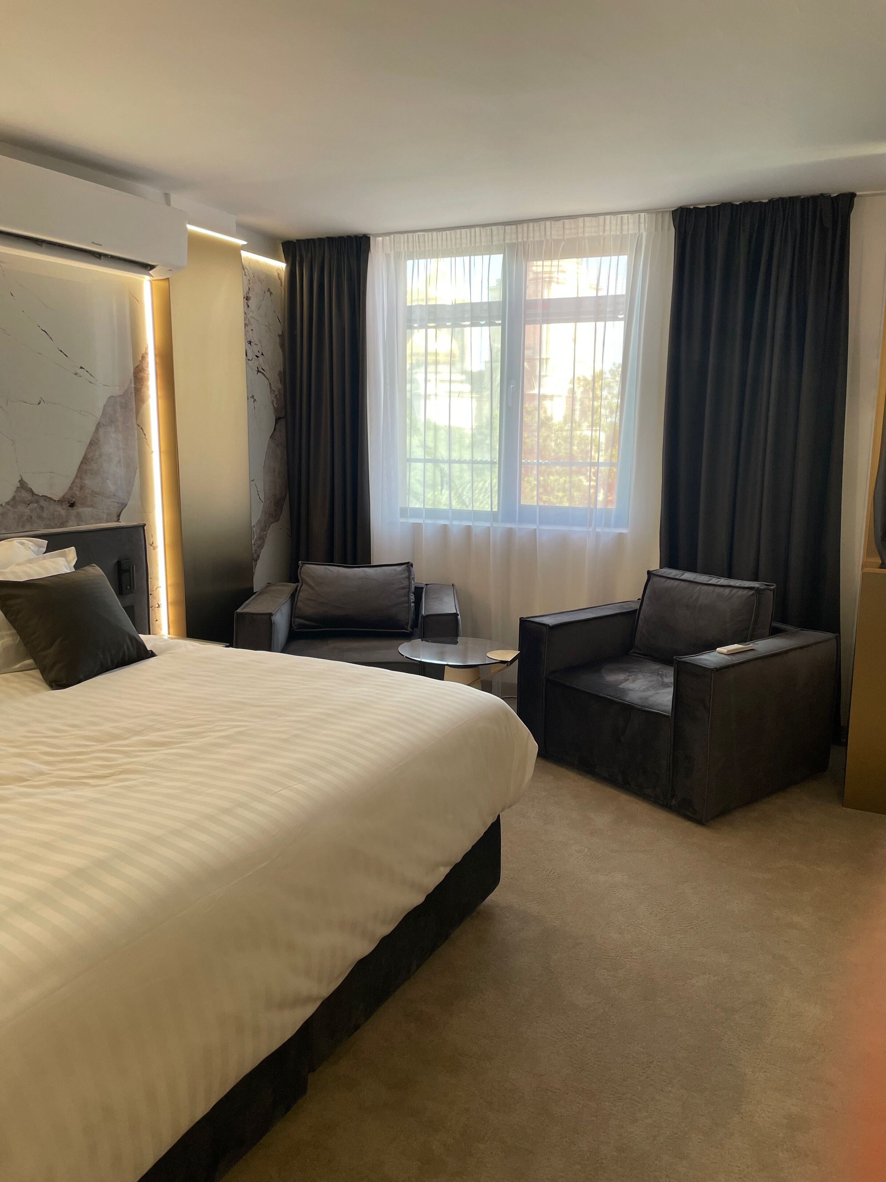 Una habitación de hotel con una cama grande, dos sillones, una mesita y un ventanal con cortinas.