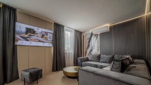 A modern hotel room with a grey sofa, a small ottoman, a round coffee table, and a large TV screen displaying a room layout. - Aslan Gold Hotel (Varna)