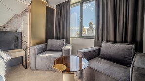 A modern hotel room with a sofa, a small round table, a desk, and a view of a building with domes. - Aslan Gold Hotel (Varna)