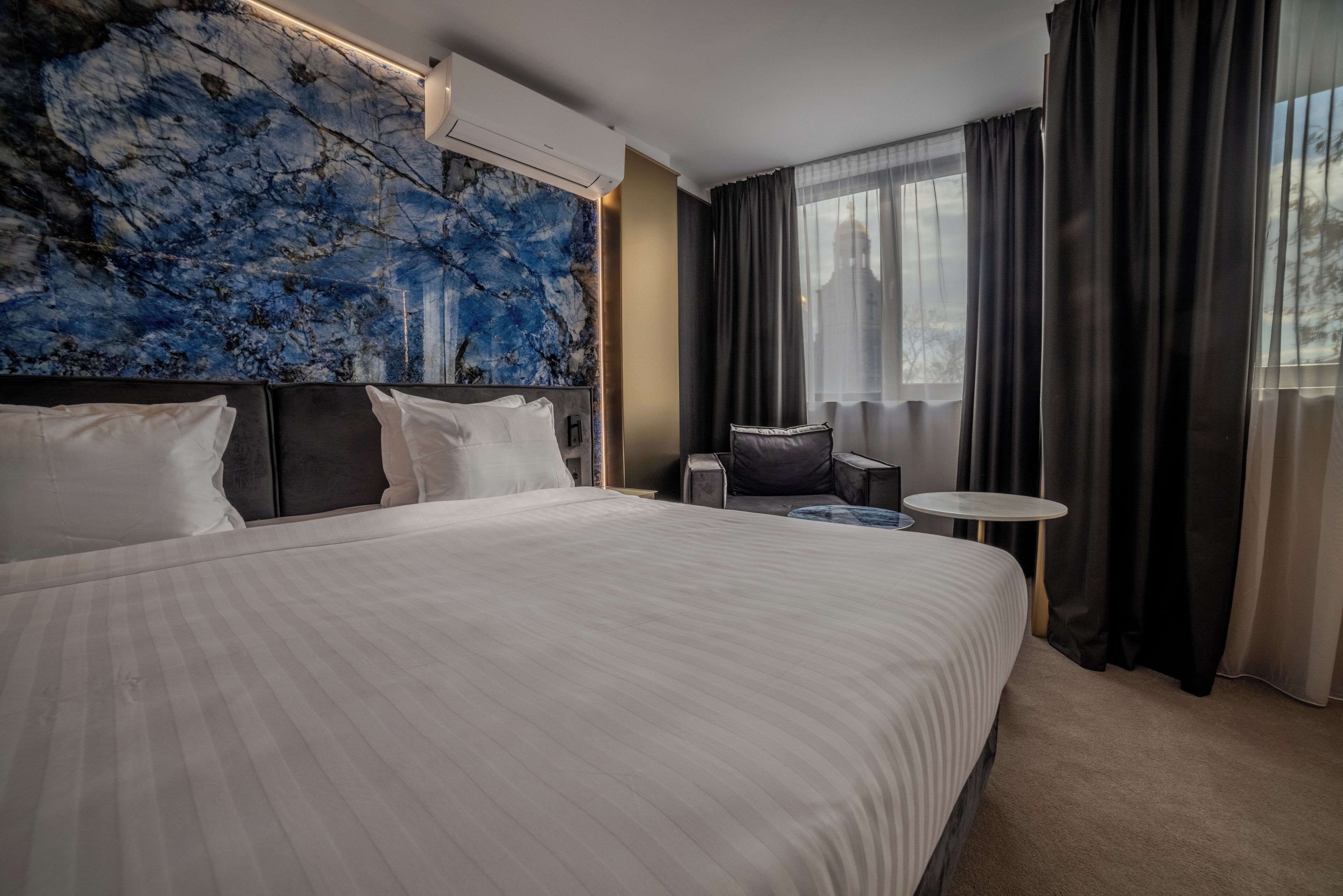 A modern hotel room with a large bed, a marble accent wall, a small round table, and a view of the city through large windows.