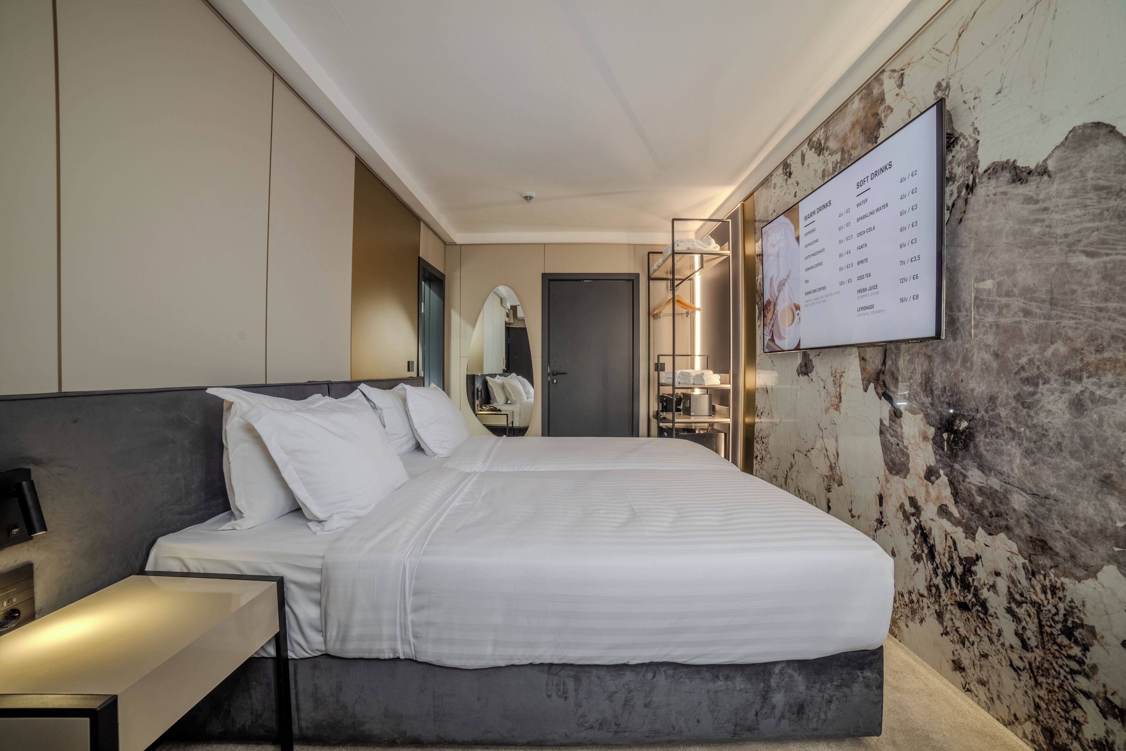 A modern hotel room with a large bed, a bedside table, a mirror, and a wall-mounted menu listing soft drinks.