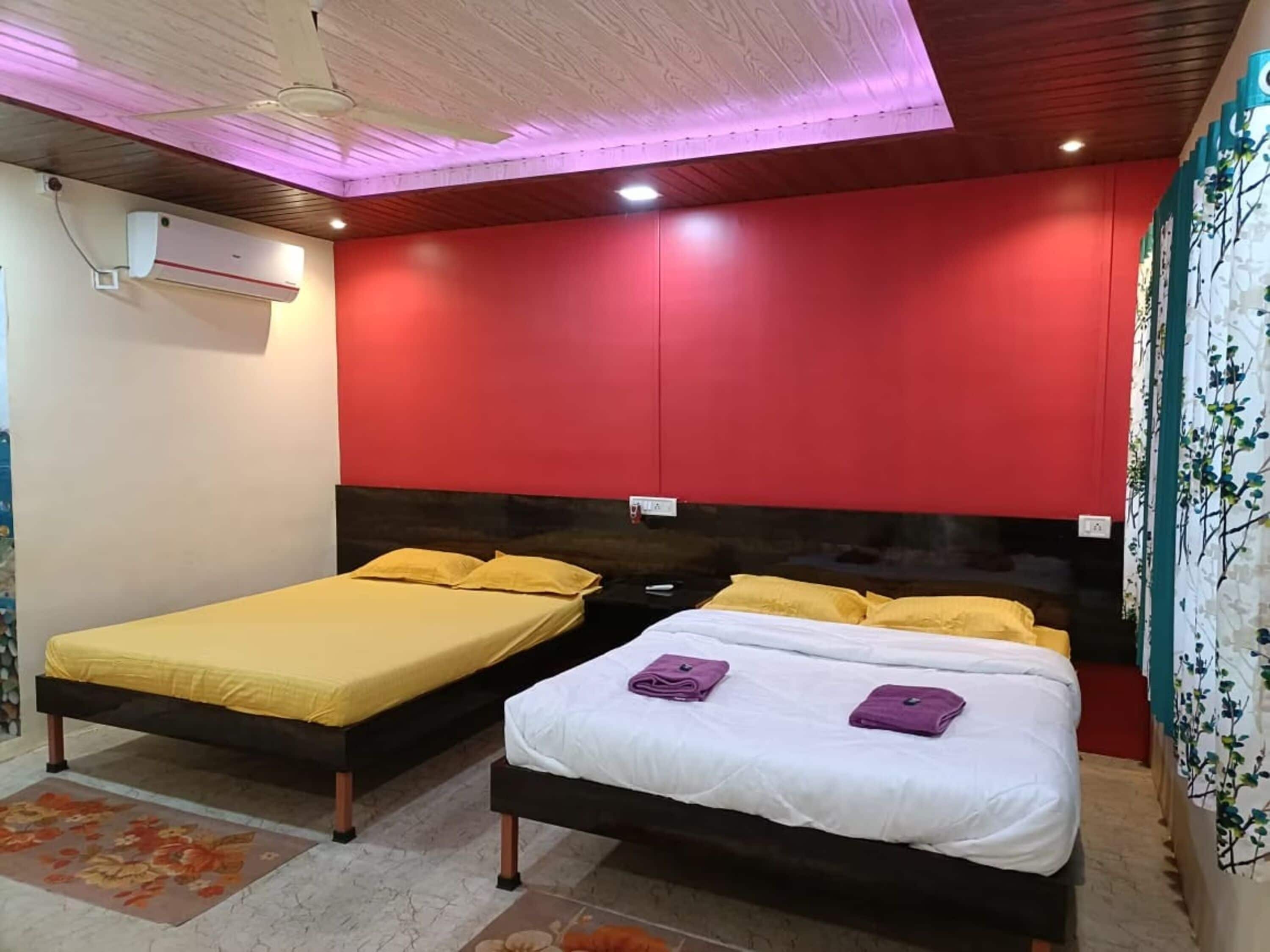 A room with two beds, one with yellow bedding and the other with white bedding, both with pillows. There is a ceiling fan, an air conditioning unit, and a wall with a floral mural.