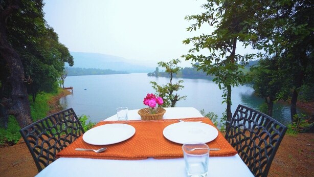 A table set for two with a view of a lake and mountains in the background. - OMKAR AGRO TOURISM AND RESORT (Mahabaleshwar)