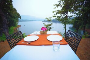 A table set for two with a view of a lake and mountains in the background. - OMKAR AGRO TOURISM AND RESORT (Mahabaleshwar)