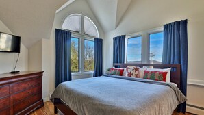 Ein Schlafzimmer mit einem großen Bett, einem Schminktisch, einem Fernseher und zwei Fenstern mit Vorhängen.