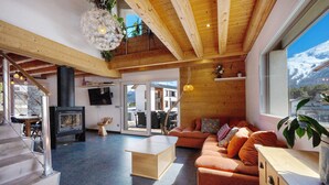 Een ruime woonkamer met een open haard, houten plafond en een groot raam met uitzicht op de bergen.