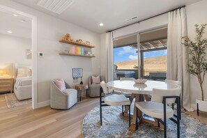 A modern living room with a round dining table, comfortable seating, a bookshelf, and a large window with a desert view. - Sandy Cheeks 6 Bedroom Townhouse (St. George)