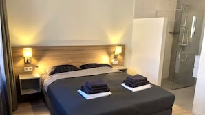 Ett hotellrum med en säng, sänglampor, en duschkabin med glasväggar och ett vitt skåp.
