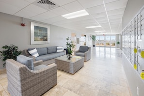 A spacious lobby with a grey sofa set, a coffee table, and a potted plant. There is a large window with a view of a landscape. - Newest 3 BR Waterfront Condo with Jaw Dropping View & Beautiful Interior! (Pensacola)