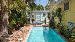 A pool area with a brick pathway, palm trees, and a small building with a window and a chair. - Old Town Studio Cottage with Full Kitchen, In-Unit Laundry & Shared Pool (Key West)