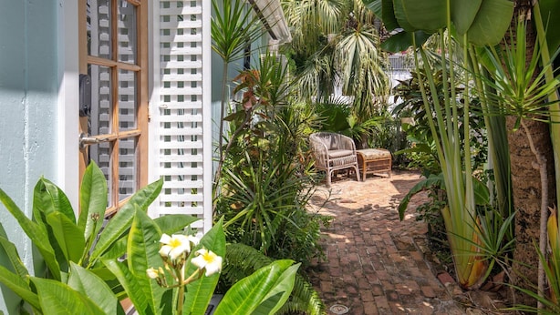 A narrow brick pathway leading to a seating area with a wicker chair and a small table, surrounded by lush tropical plants and a white lattice fence. - Old Town Studio Cottage with Full Kitchen, In-Unit Laundry & Shared Pool (Key West)