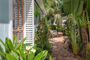 A narrow brick pathway leading to a seating area with a wicker chair and a small table, surrounded by lush tropical plants and a white lattice fence. - Old Town Studio Cottage with Full Kitchen, In-Unit Laundry & Shared Pool (Key West)
