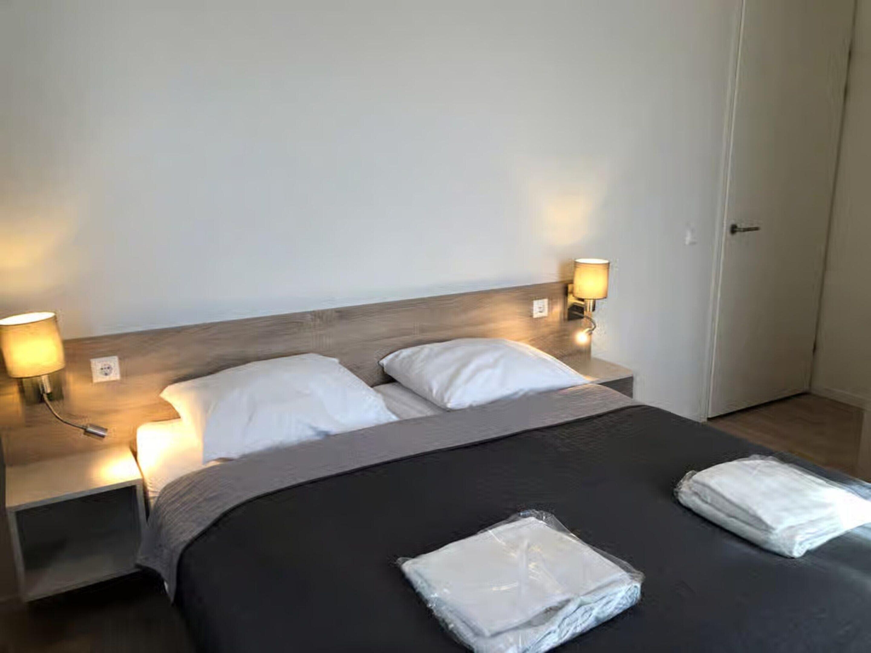 Cama bem arrumada com almofadas brancas e um edredom preto, duas luminárias de cabeceira e uma porta fechada.