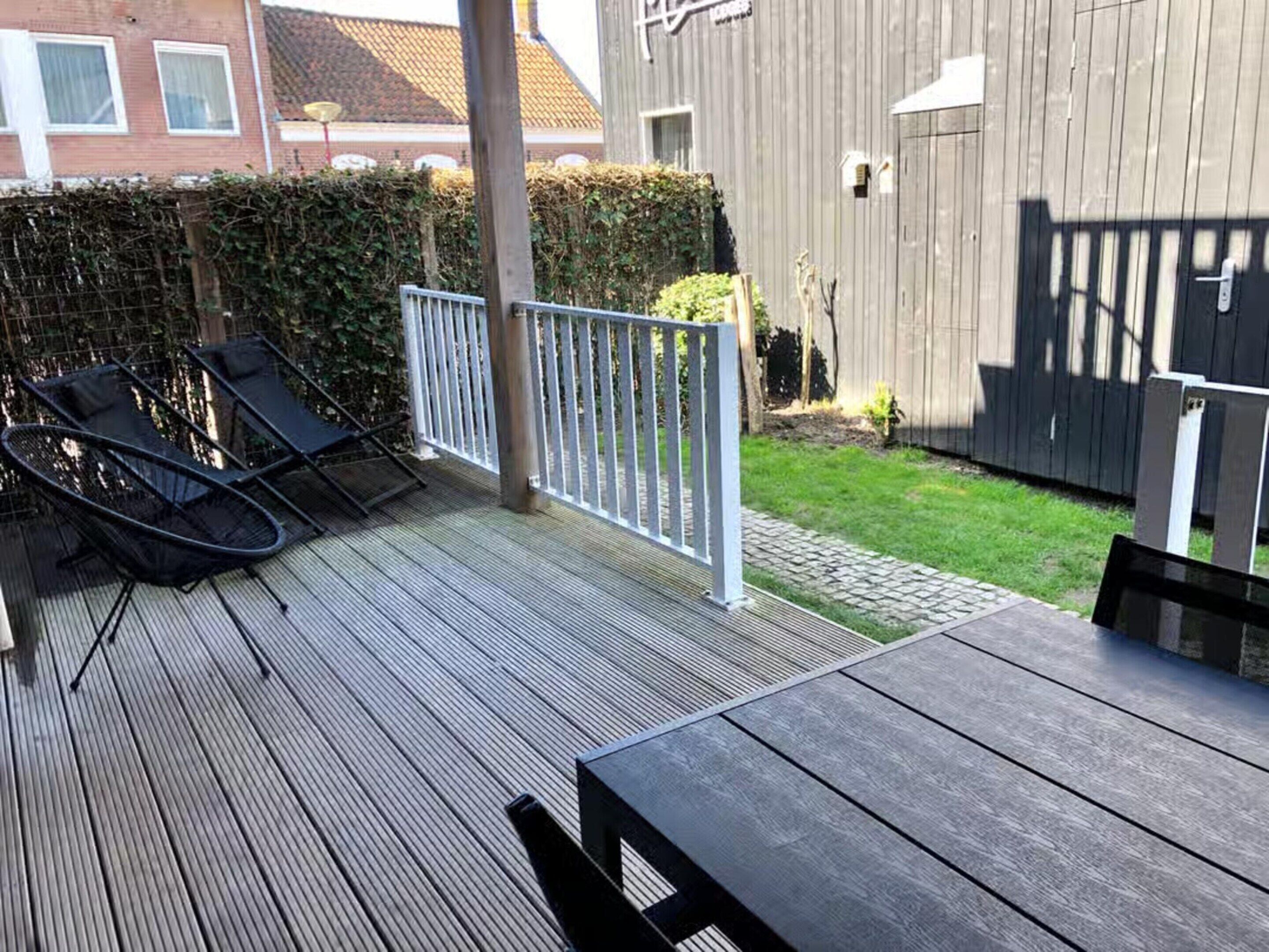 A wooden deck with a table and chairs, a white fence, and a hedge.