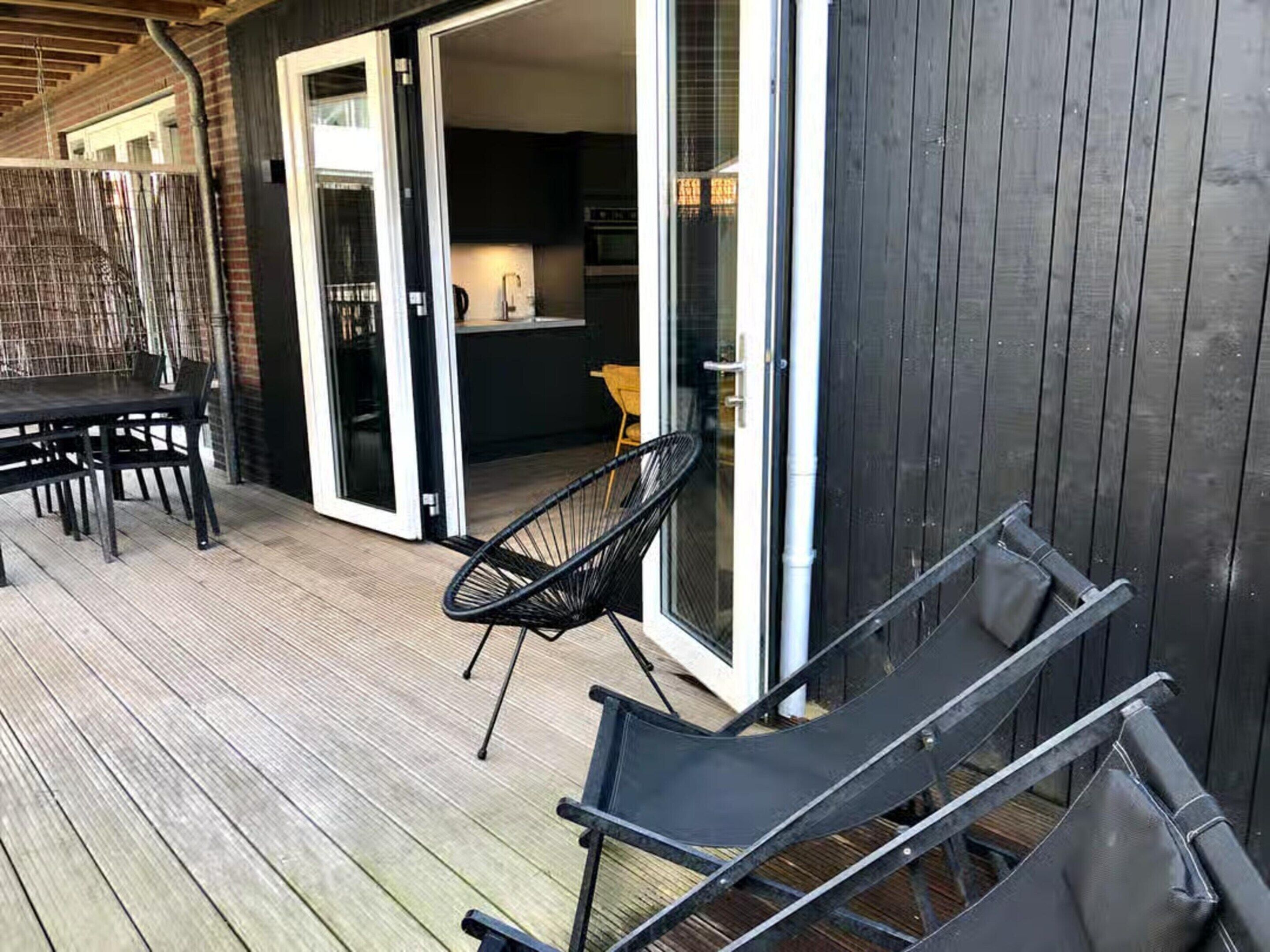 Een houten terras met een tafel en stoelen, een schuifdeur naar een keuken, en een donkere buitenmuur.