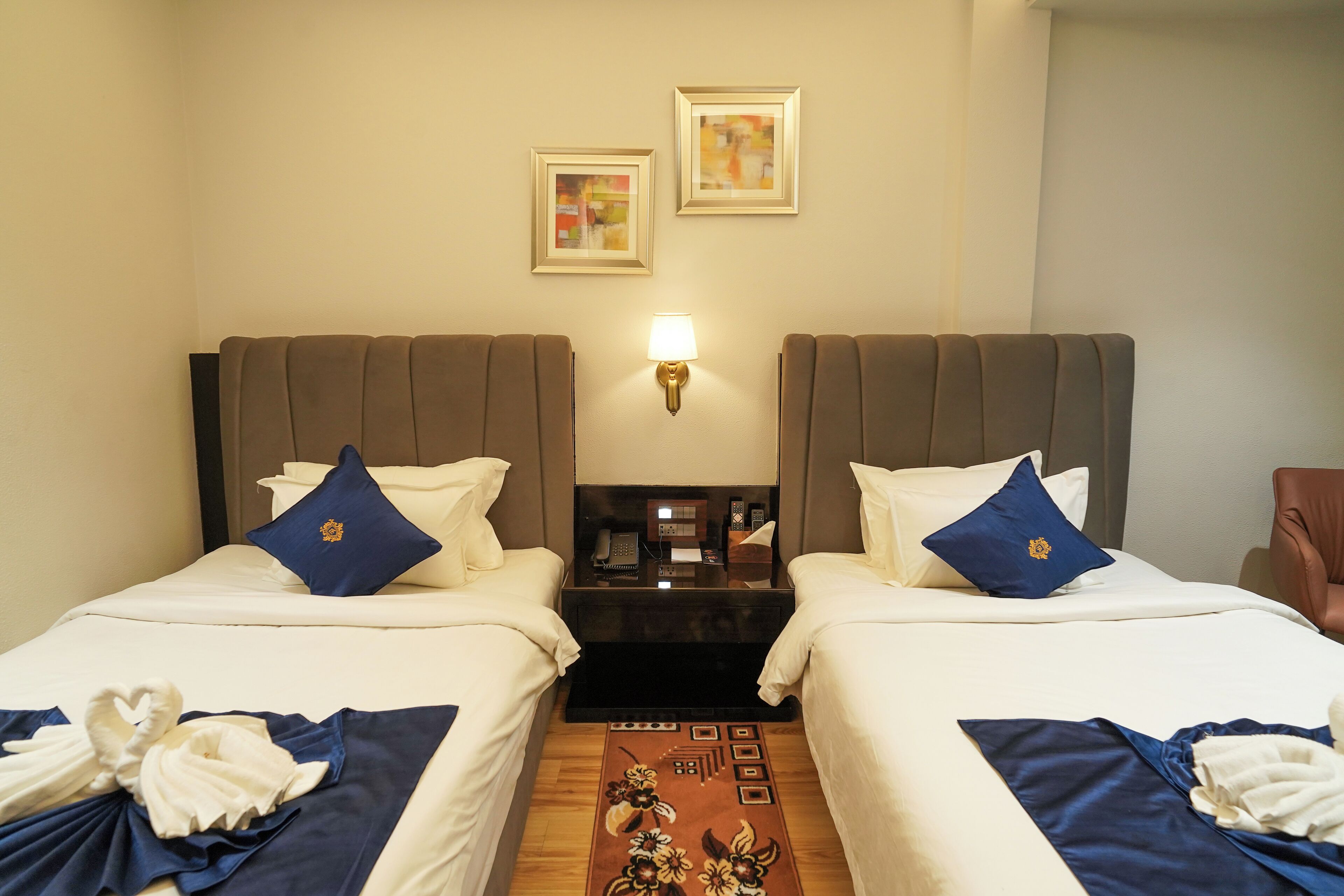 A hotel room with two beds, each with a pillow and a blanket. There is a nightstand with a lamp and a phone between the beds.