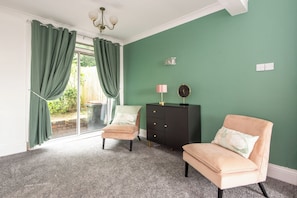 A room with a green wall, a black cabinet, two chairs, a lamp, and a window with curtains. - Toast Stays - The Garnet Rest (Brandon)