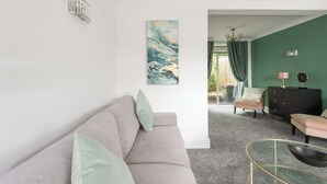 A modern living room with a beige sofa, a glass coffee table, and a green wall with a painting. - Red Courts (Brandon)