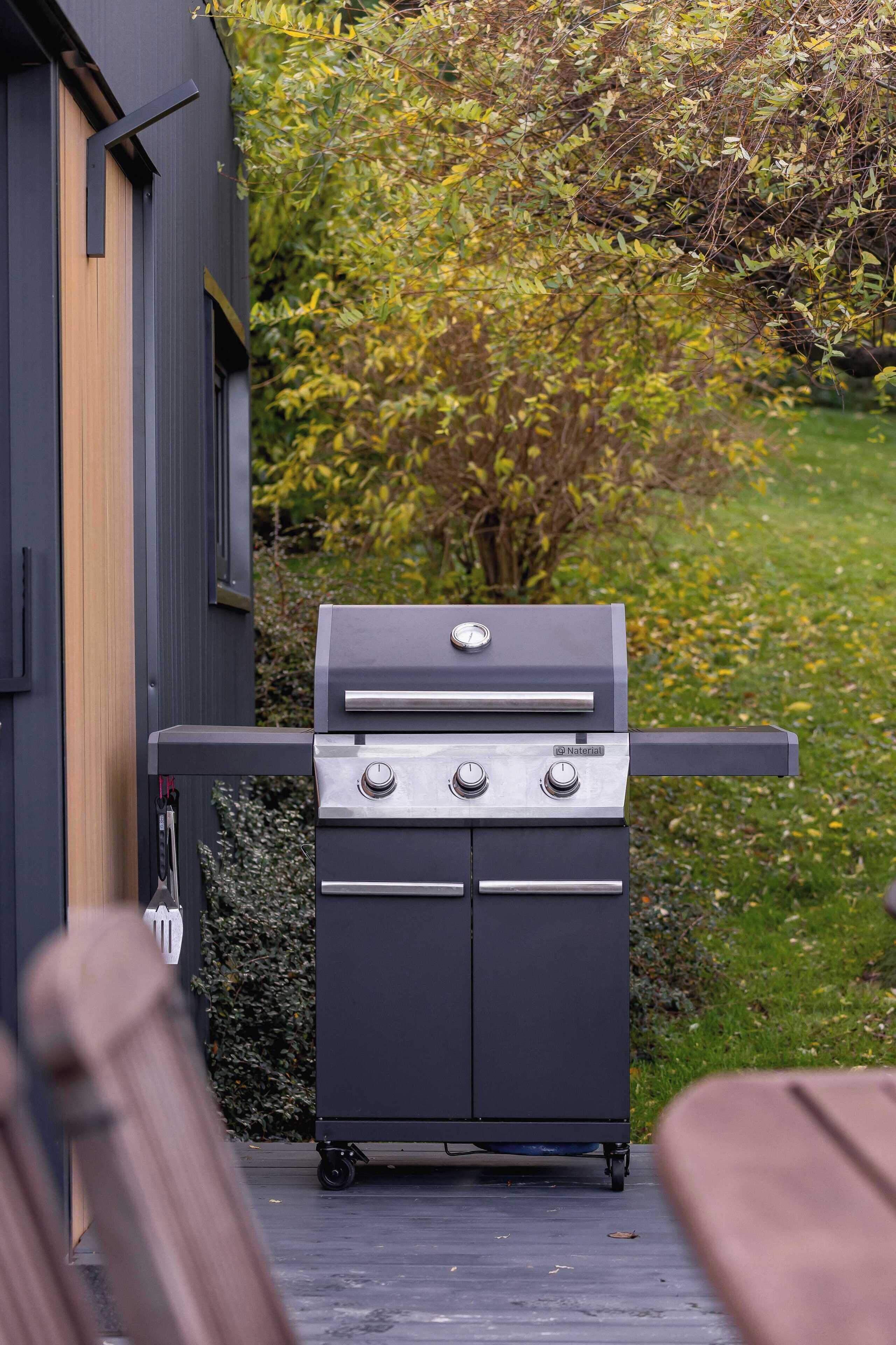 Una griglia barbecue portatile con quattro fuochi, posizionata su un patio con un pavimento in legno e un giardino sullo sfondo.