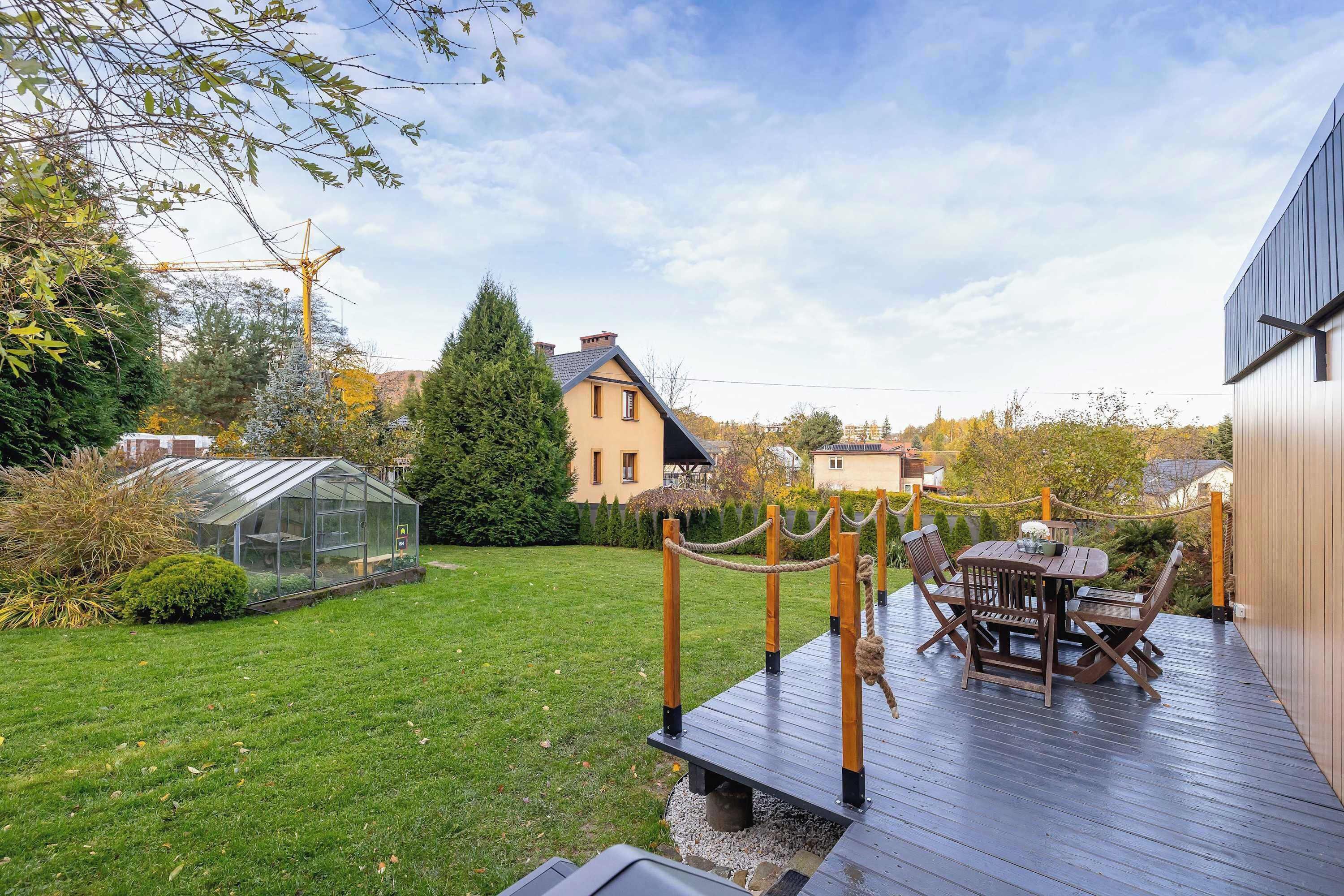 Una terraza de madera con una mesa y sillas, rodeada de vegetación y una casa al fondo.