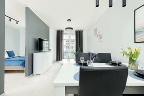 A modern hotel room with a bed, a TV, a dining area with a table and chairs, and a view of the city through large windows. - Comfortable Apartment | Sauna, Gym and Parking (Kołobrzeg)