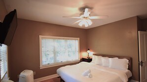 A bedroom with a bed, a ceiling fan, a heater, a window with blinds, and a nightstand with a lamp.