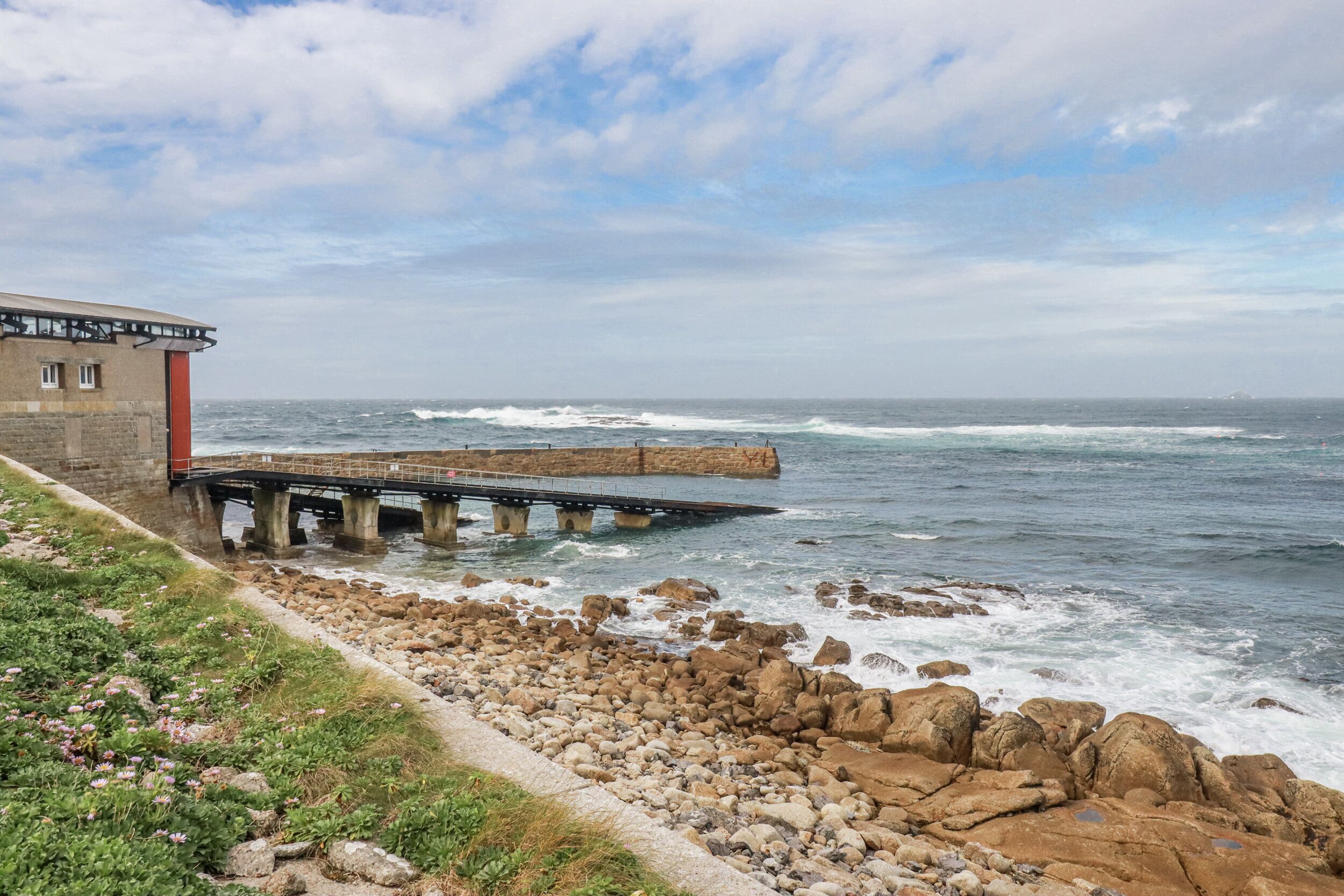 A coastal scene with a stone building, a pier extending into the sea, and a rocky shoreline with vegetation.