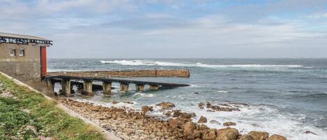 A coastal scene with a stone building, a pier extending into the sea, and a rocky shoreline with vegetation.