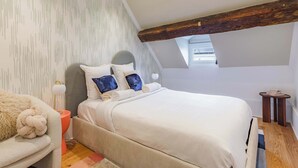 A bedroom with a sloped ceiling, a bed with white bedding, a grey sofa, a small round table, and a patterned wall. - 11 Beaumarchais 5D (Paris)